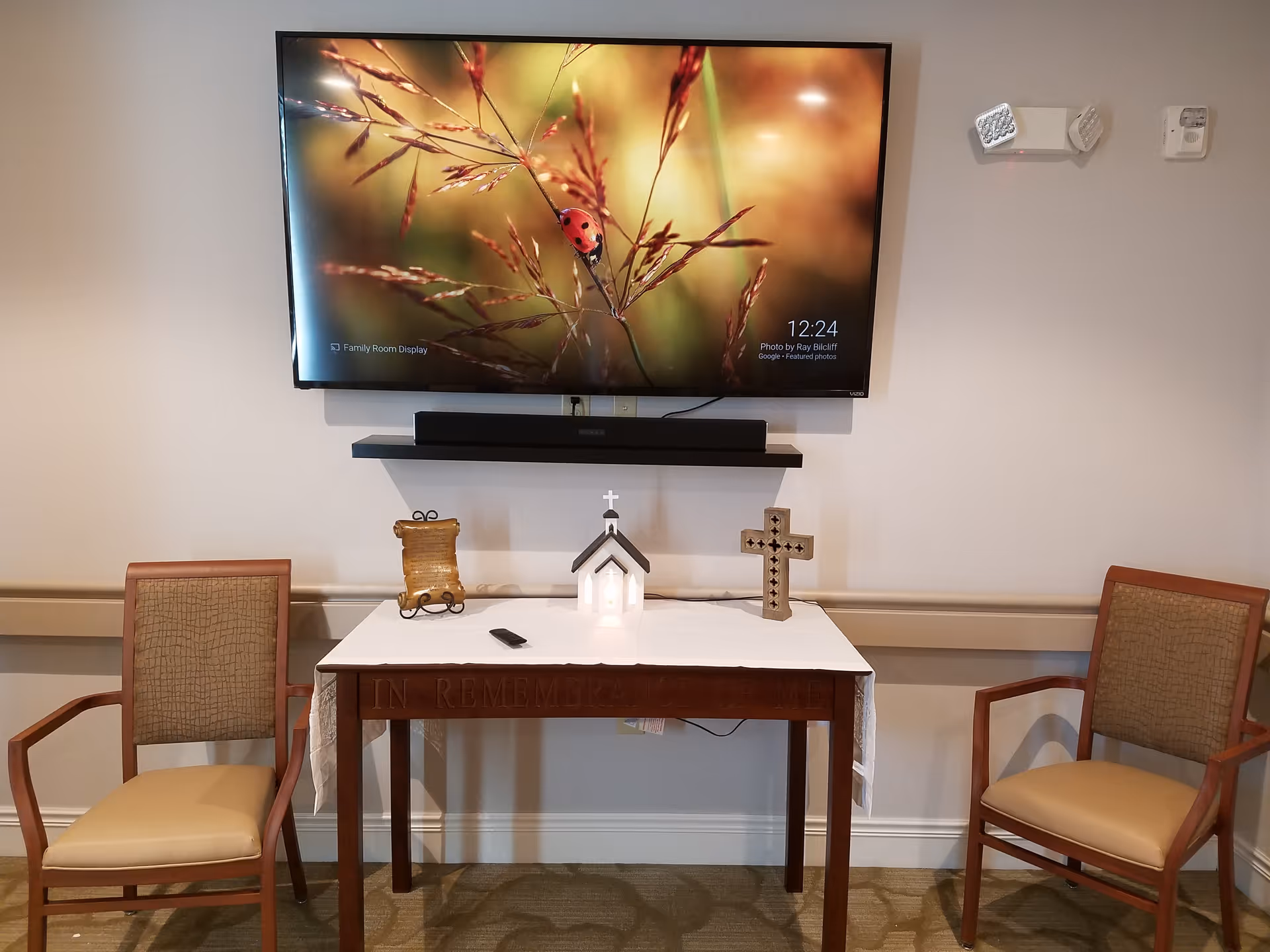 A small memorial table with a white cloth, a miniature church with a cross, a decorative cross, and a scroll on it, flanked by two wooden chairs with beige cushions. Above the table is a wall-mounted flat-screen TV displaying a close-up image of a ladybug on grass. The wall is light-colored with a mounted speaker and a smoke detector.