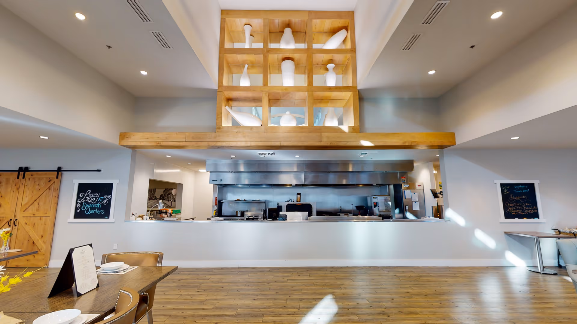 Interior view of a dining area with a large open kitchen in the background. The kitchen has stainless steel appliances and a wooden decorative shelving unit above it holding white vases. The dining area has wooden floors, tables set with plates and napkins, and two chalkboards on the walls. One chalkboard reads 'Legacy Savannah Quarters'.