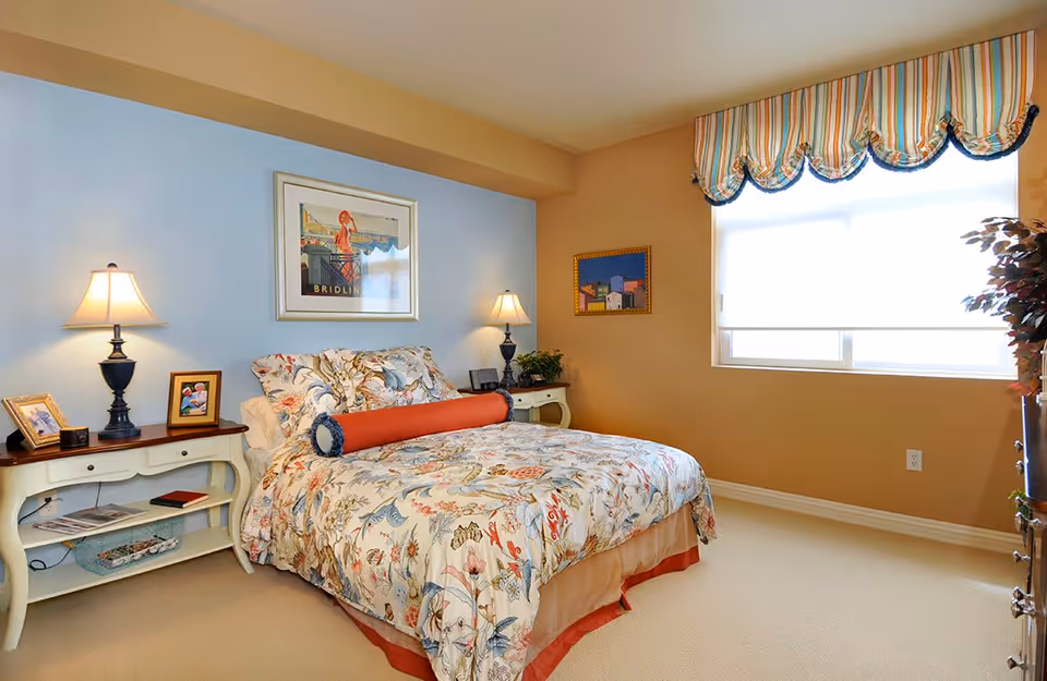 A cozy bedroom with a floral-patterned bedspread and a cylindrical orange pillow on the bed. There are two nightstands on either side of the bed, each with a lamp and framed photos. The walls are painted in light blue and beige tones, with two framed pictures hanging. A window with a striped valance lets in natural light, and a plant is placed near the window.