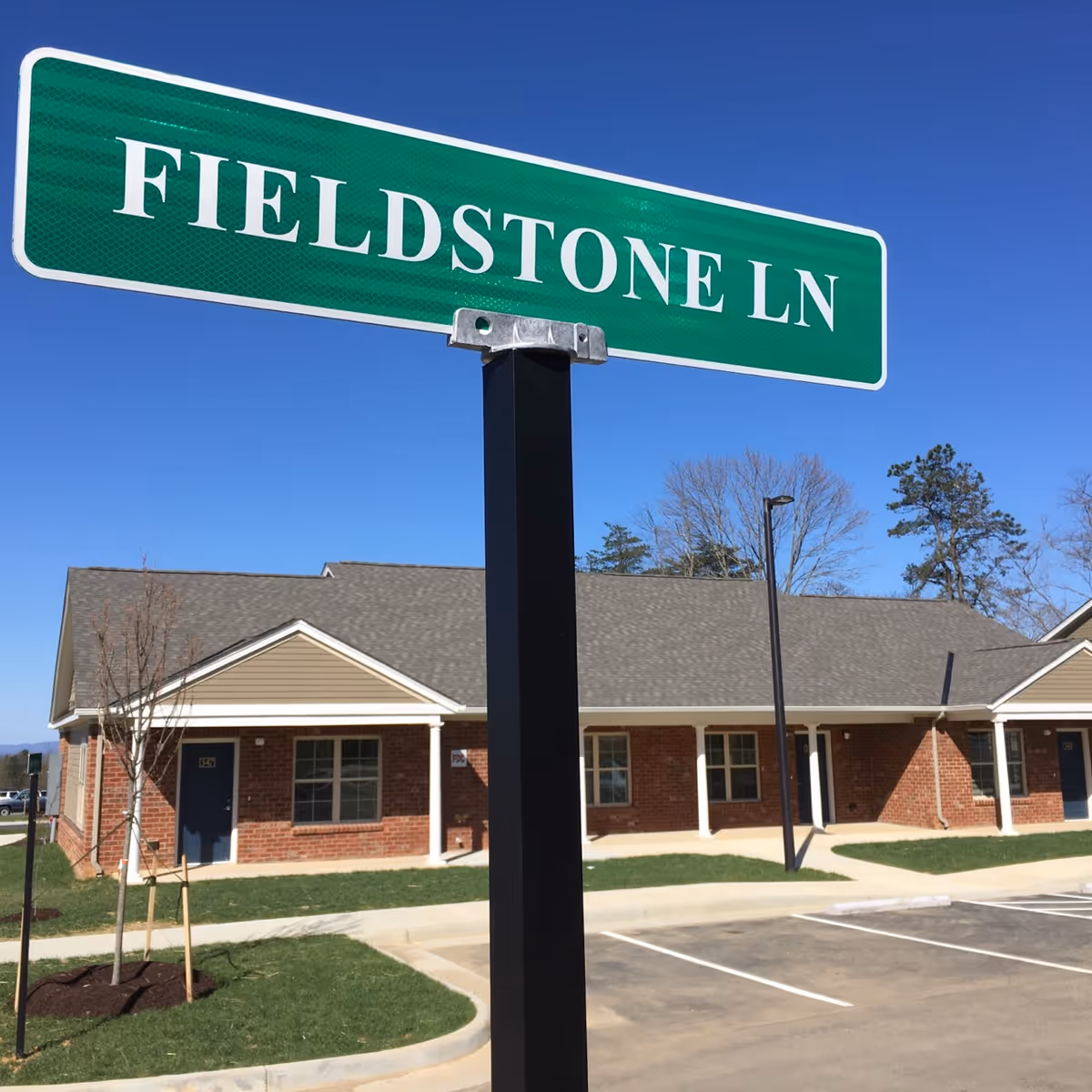 Green street sign reading 'FIELDSTONE LN' in front of a single-story brick building with a gray roof, windows, and blue doors, under a clear blue sky.
