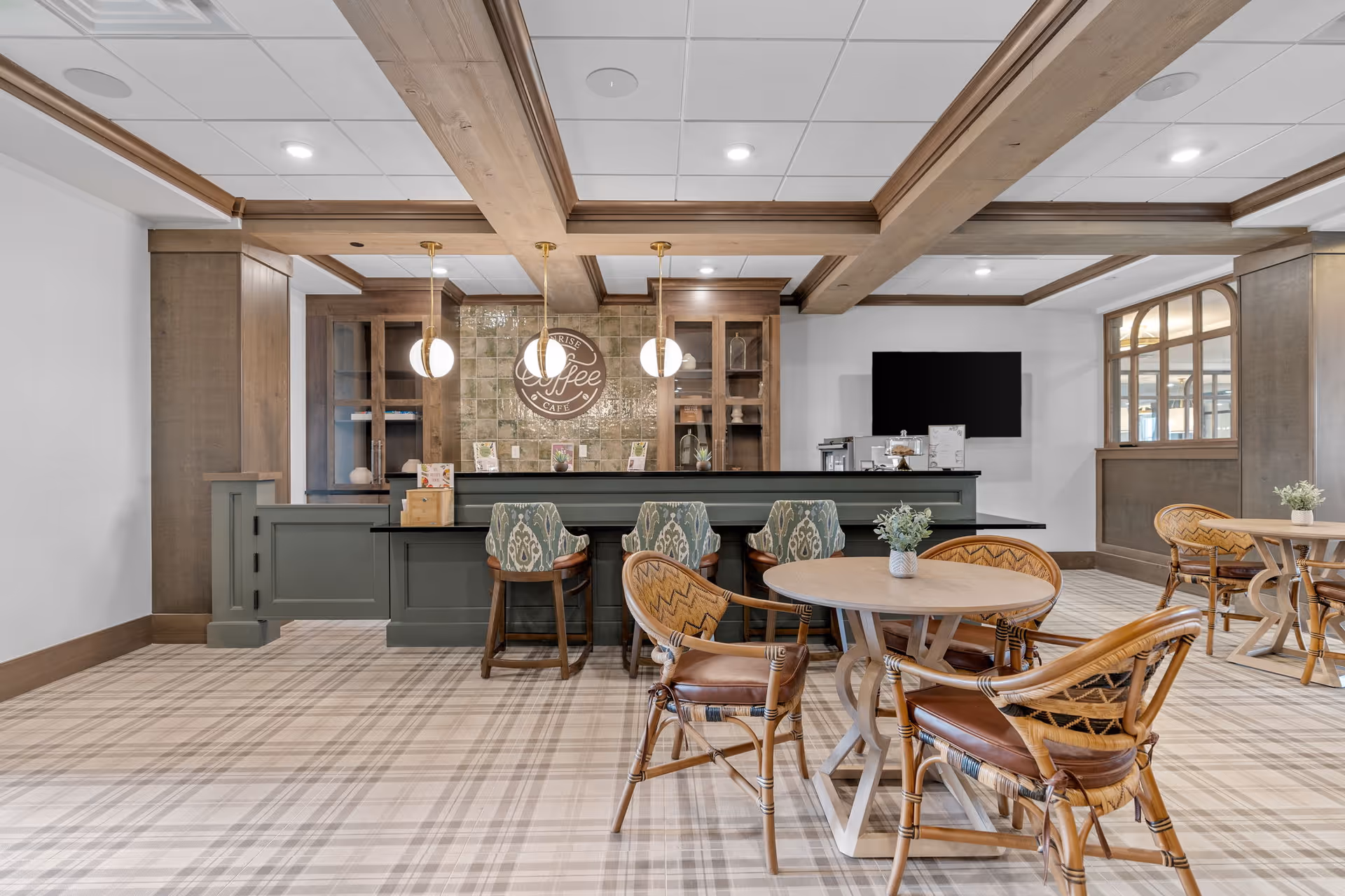 A cozy coffee cafe area inside a senior living facility featuring a counter with three patterned bar stools, round tables with wicker chairs, wooden beams on the ceiling, and a sign on the tiled wall that reads 'Coffee Cafe'. The space has a warm, inviting atmosphere with soft lighting and a plaid carpeted floor.