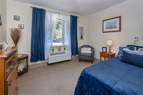 A cozy bedroom with a bed covered in blue bedding, a gray armchair with a pillow, a wooden nightstand with a lamp, a framed picture on the wall, a window with blue curtains and sheer white drapes, and a wooden dresser with decorative items.