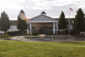 Front exterior view of a single-story building with a covered entrance supported by white columns. There are trees and shrubs around the entrance, a flagpole with an American flag on the right side, and a parking area with marked spaces in front. The sky is partly cloudy.