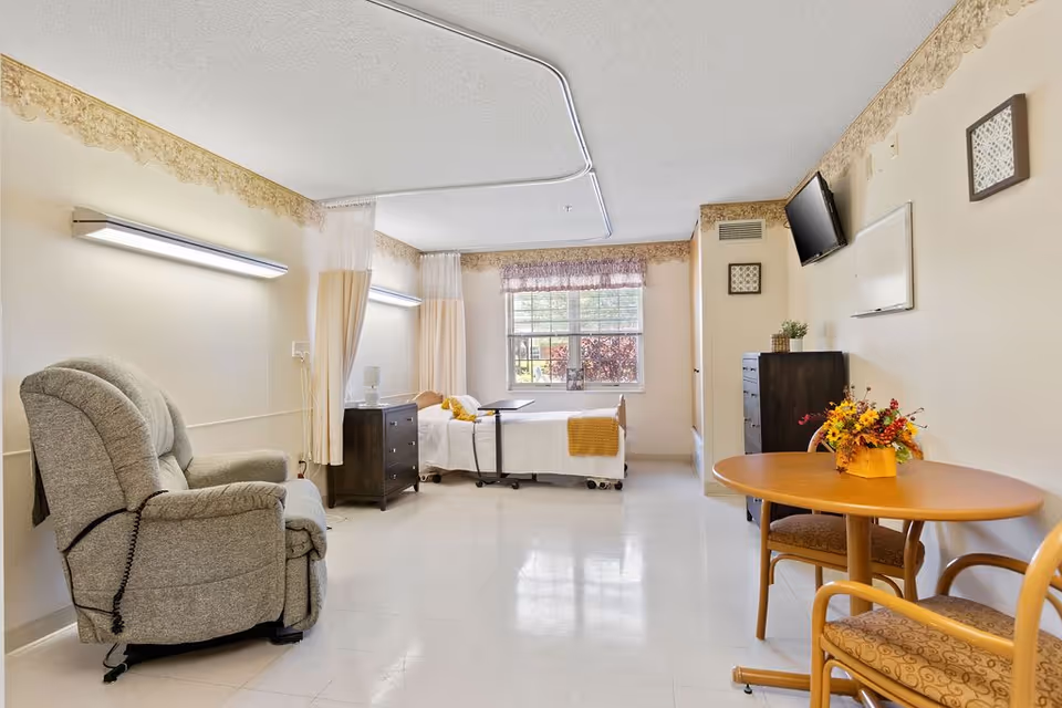 A bright nursing facility room with a hospital bed near a window, a gray recliner chair, a wooden table with two chairs, a wall-mounted TV, and light fixtures on the walls. The room has light-colored walls with a decorative border near the ceiling and a clean tiled floor.