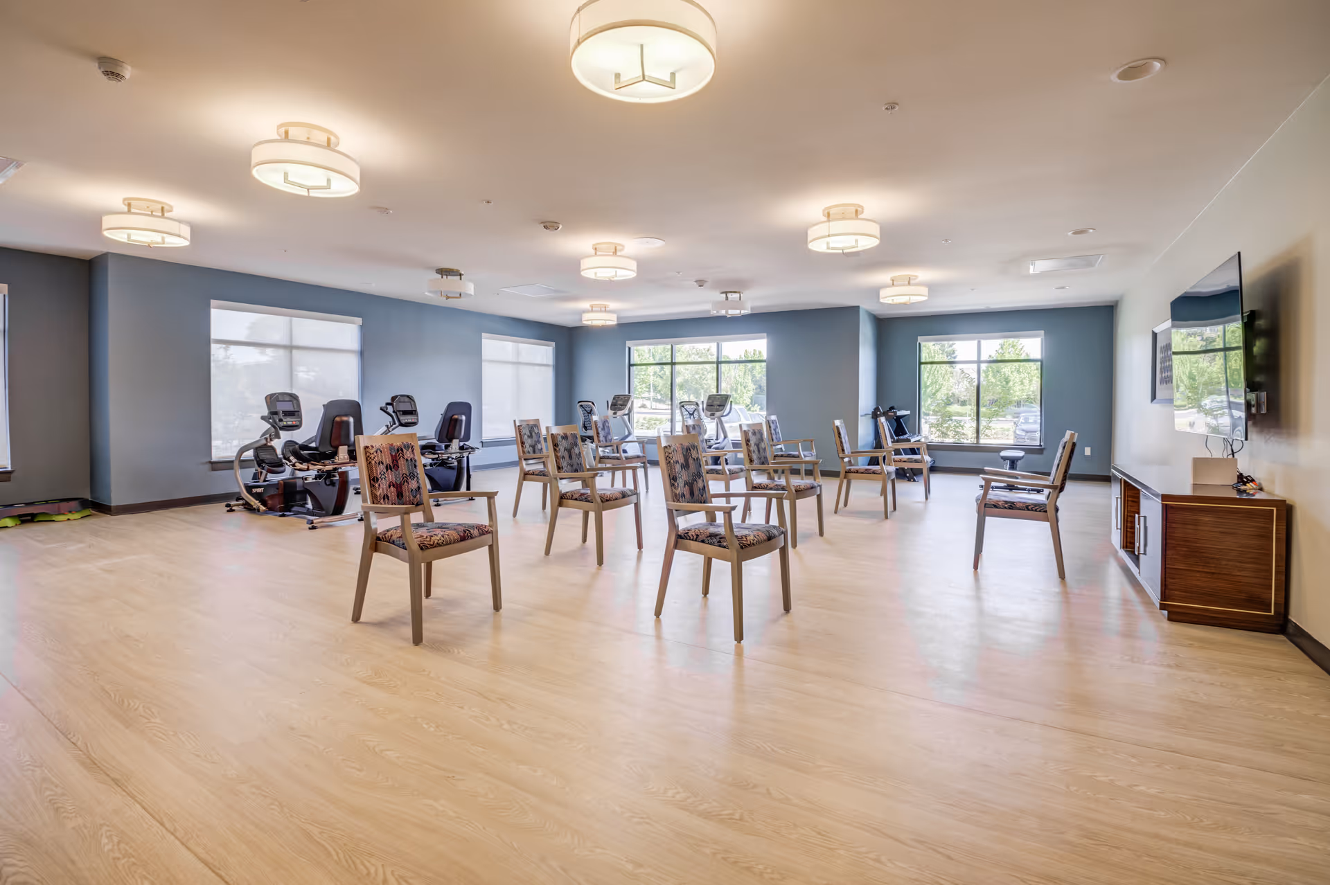 A spacious exercise room with wooden flooring and blue walls, featuring several stationary exercise bikes near the windows and multiple chairs arranged in rows facing a wall-mounted TV. The room is well-lit with ceiling lights and natural light from large windows.