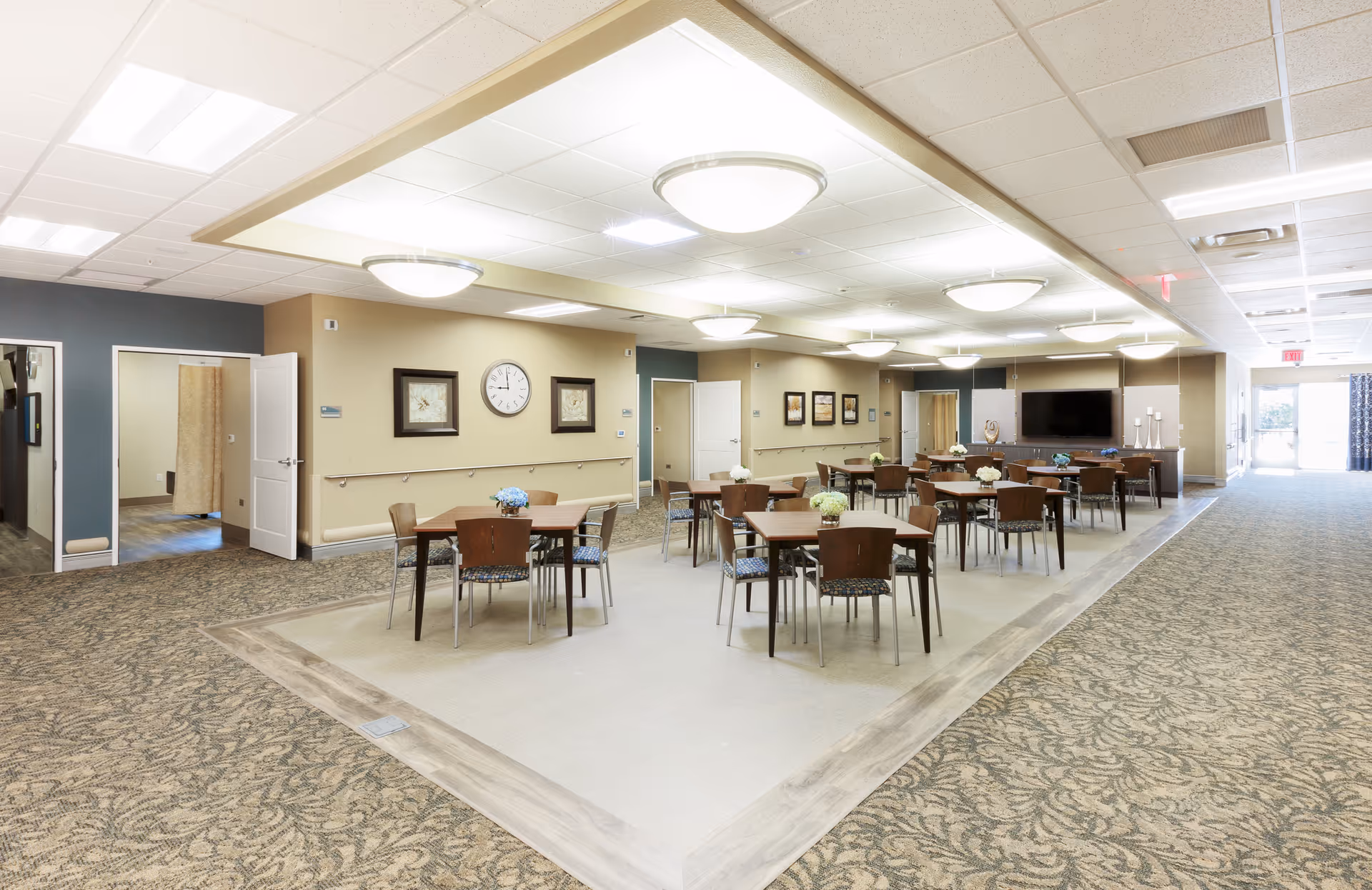 A spacious dining area in a senior living facility with several square wooden tables and chairs arranged neatly. The room has a beige and blue color scheme with framed artwork and a clock on the walls. Ceiling lights provide bright illumination, and there is a large flat-screen TV mounted on the far wall. The floor is carpeted with a patterned design, except for the dining area which has a different flooring. Doors and hallways lead to other parts of the facility.