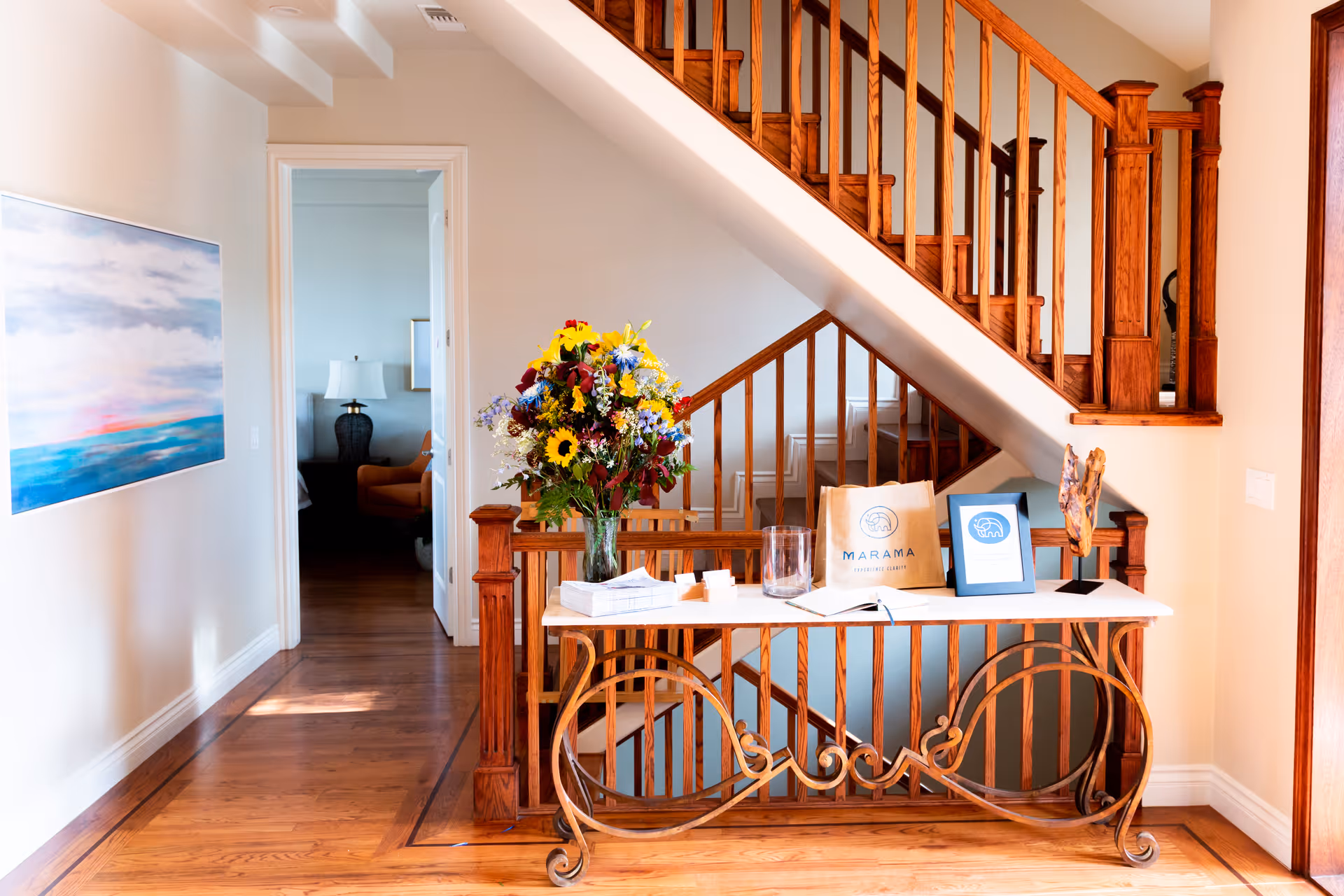 A bright interior hallway with wooden flooring and a staircase with wooden railings. A decorative table with a vase of colorful flowers, a paper bag, and framed items is placed under the staircase. A doorway leads to another room with a lamp and chair visible. A large painting with blue and pink hues hangs on the left wall.