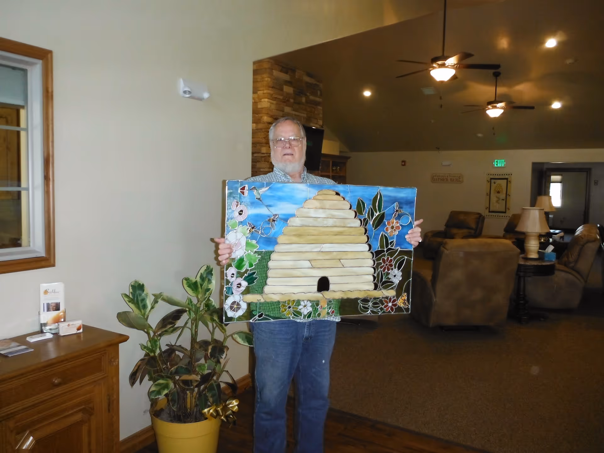 An elderly man standing indoors holding a colorful painting of a beehive surrounded by flowers and bees. The room has beige walls, ceiling fans with lights, comfortable armchairs, a wooden cabinet with brochures, and a potted plant nearby.