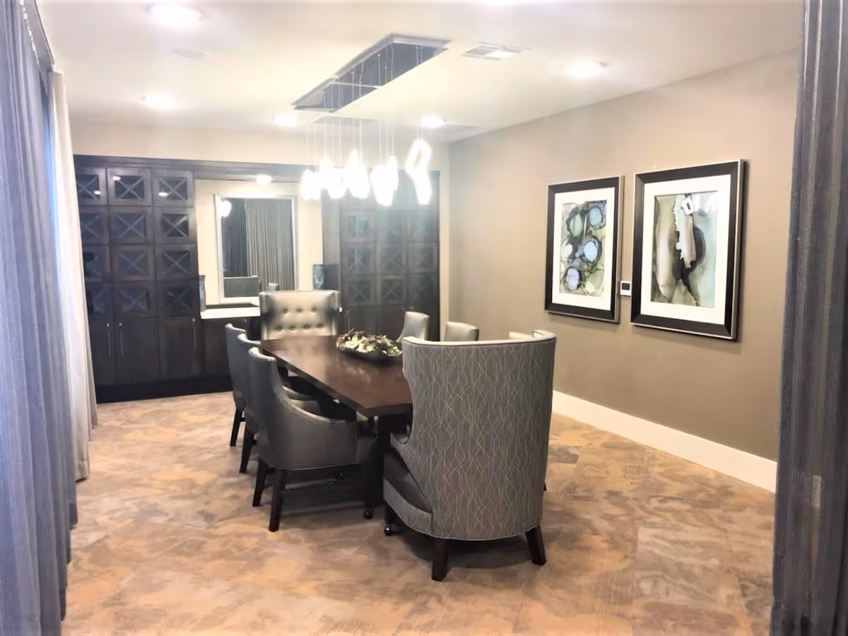 A modern dining room with a long dark wooden table surrounded by eight upholstered chairs, two of which have high backs. The room features a beige carpet, neutral-colored walls with two abstract framed artworks, a large mirror on the far wall, and built-in dark wood cabinetry. A contemporary chandelier with multiple hanging lights illuminates the space.