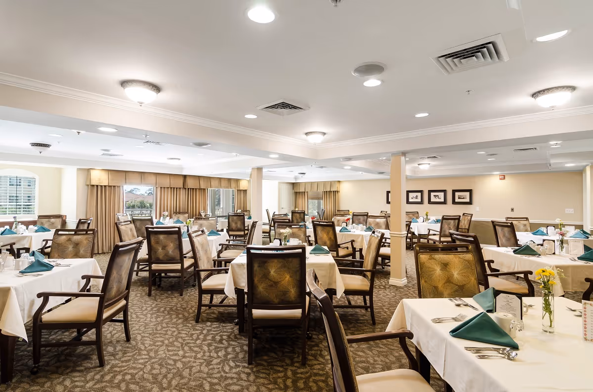 A spacious dining room with multiple tables covered in white tablecloths, each set with teal folded napkins, silverware, and small flower vases. The room has beige walls, carpeted floors, and large windows with beige curtains allowing natural light to enter.