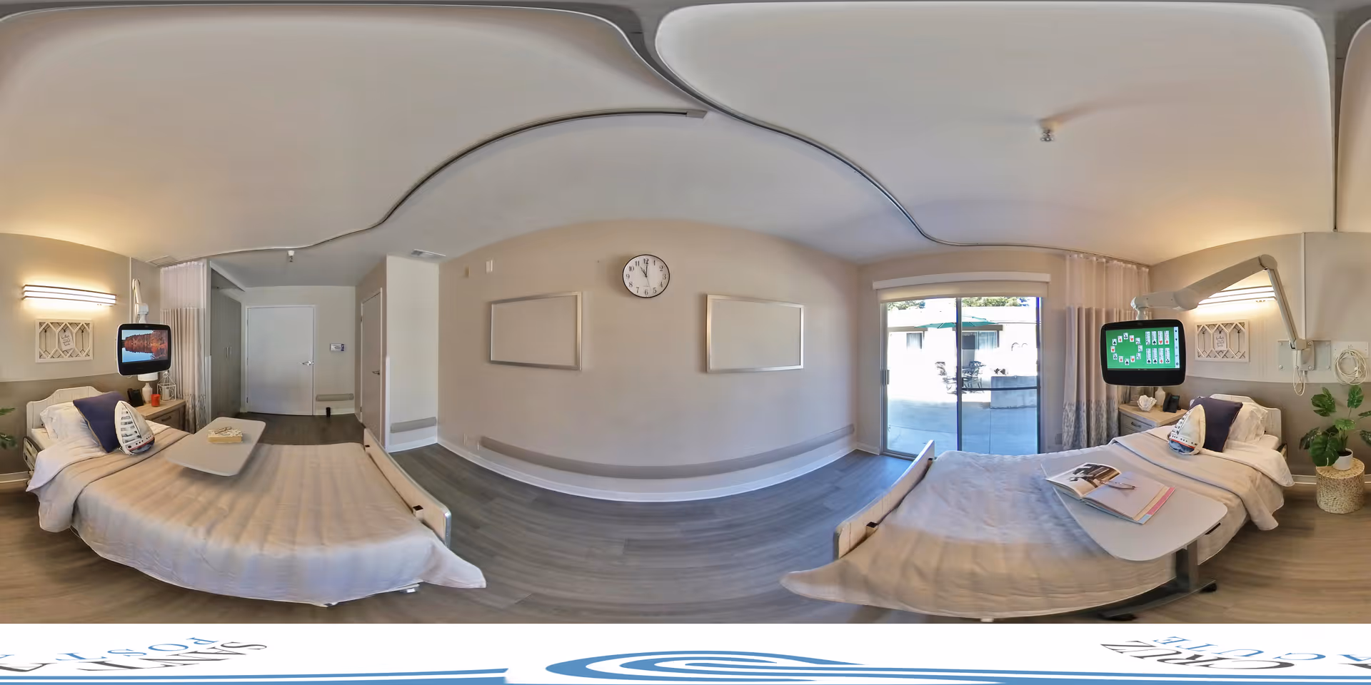 Wide-angle view of a bright two-bed patient room with hospital beds, overbed tables, wall-mounted monitors, a clock, and sliding glass doors to an outdoor patio.