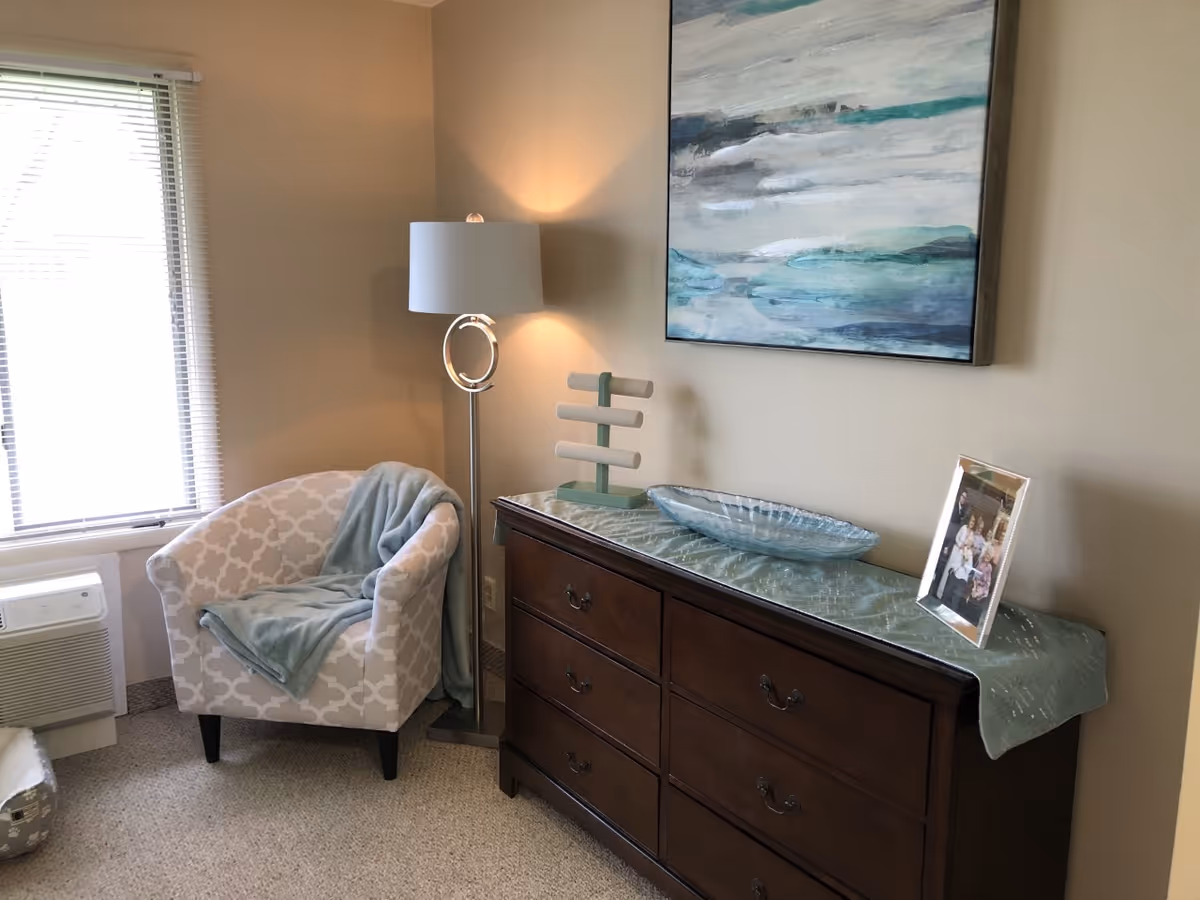 A cozy corner of a room featuring a patterned armchair with a light blue blanket draped over it, a modern floor lamp with a circular design, and a dark wooden dresser topped with a light blue runner, a decorative glass bowl, a jewelry stand, and a framed family photo. A large abstract painting with blue and gray tones hangs on the beige wall above the dresser. A window with blinds is visible to the left.