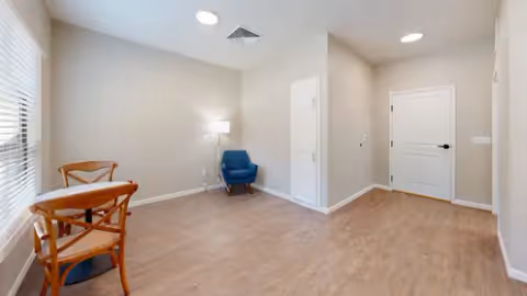 A small, minimally furnished room with light wood flooring and beige walls. The room contains a round wooden table with two wooden chairs near a window with blinds, and a blue upholstered armchair in the corner next to a floor lamp. There are two white doors on the far wall and recessed ceiling lights.