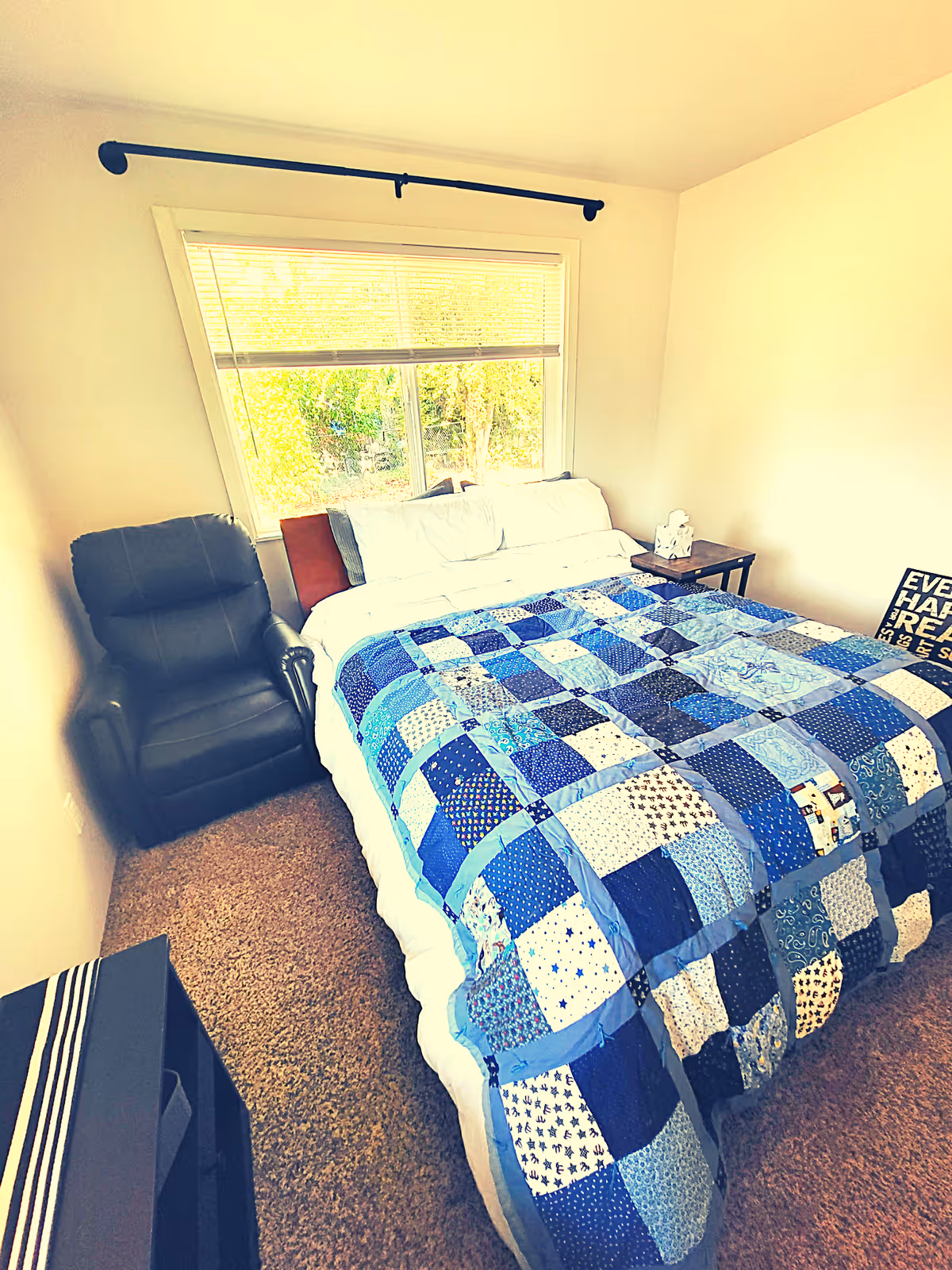A bedroom with a large window letting in natural light. The room features a bed with a blue patchwork quilt, white pillows, and a wooden headboard. To the left of the bed is a black recliner chair, and to the right is a small wooden side table with a tissue box on it. The floor is carpeted in a light brown color.