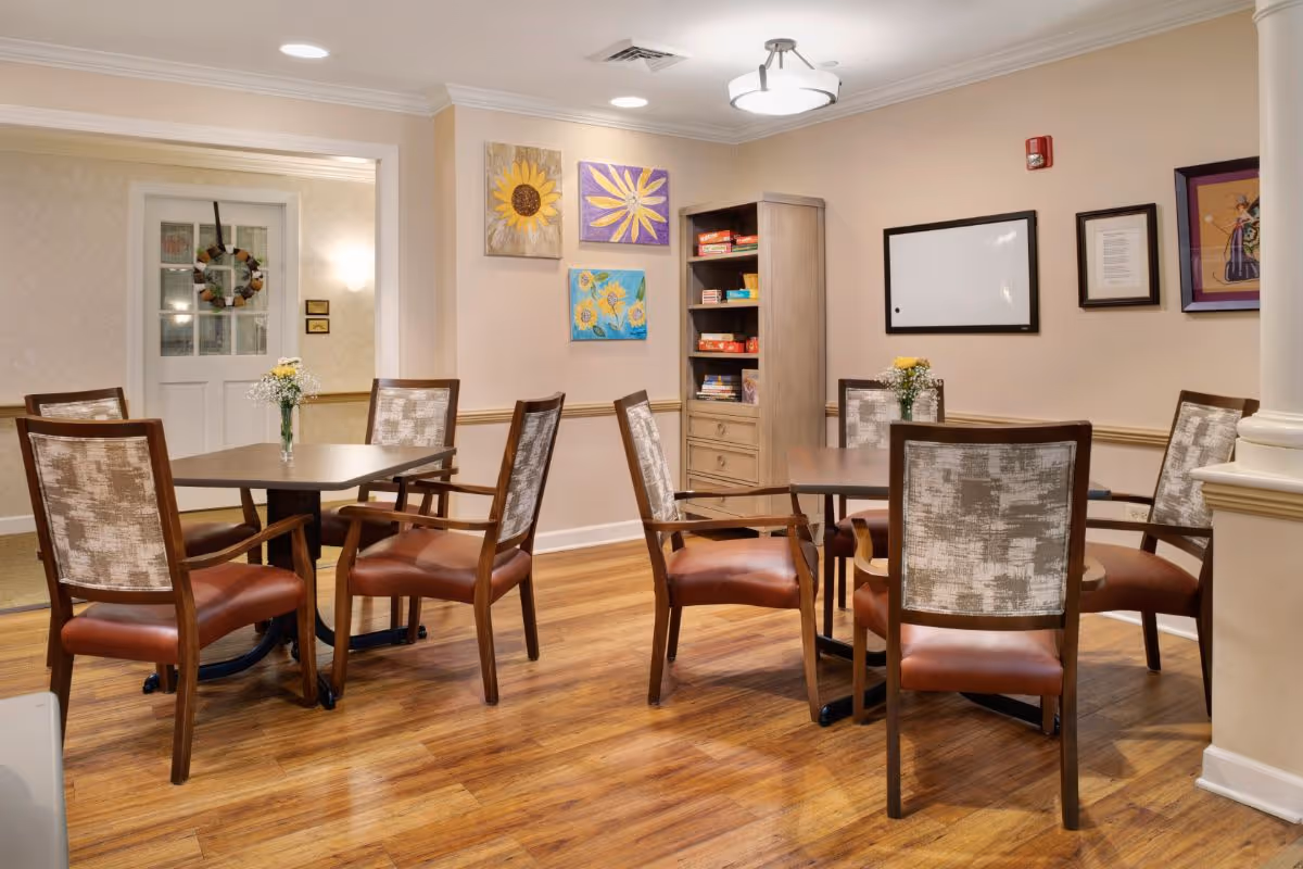 A cozy interior room with two square tables, each surrounded by four wooden chairs with cushioned seats and backs. The room has wooden flooring, beige walls adorned with colorful sunflower paintings, a whiteboard, and framed pictures. A wooden cabinet with board games is visible in the corner, and small vases with flowers are placed on the tables.