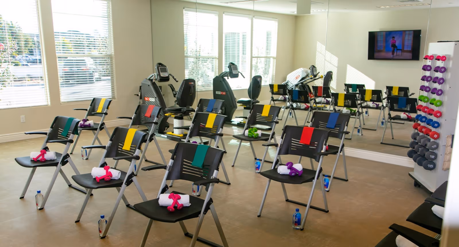 A fitness room with multiple black chairs arranged in rows, each chair equipped with a colored resistance band, a pair of small dumbbells, a rolled white towel, and a bottle of water on the floor beside it. The room has large windows with blinds, exercise bikes, a wall-mounted TV showing a workout video, and a rack of various colored dumbbells against a mirrored wall.