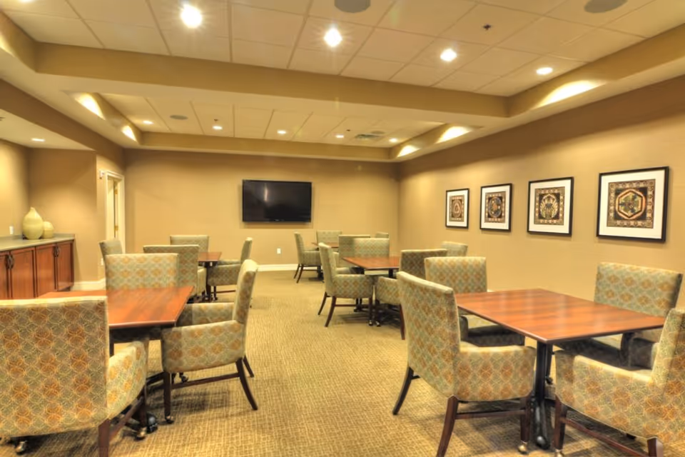 A well-lit room with multiple wooden tables and cushioned chairs arranged neatly. The walls are beige with framed artwork hanging on one side. A flat-screen TV is mounted on the far wall. The ceiling has recessed lighting and a drop ceiling design.