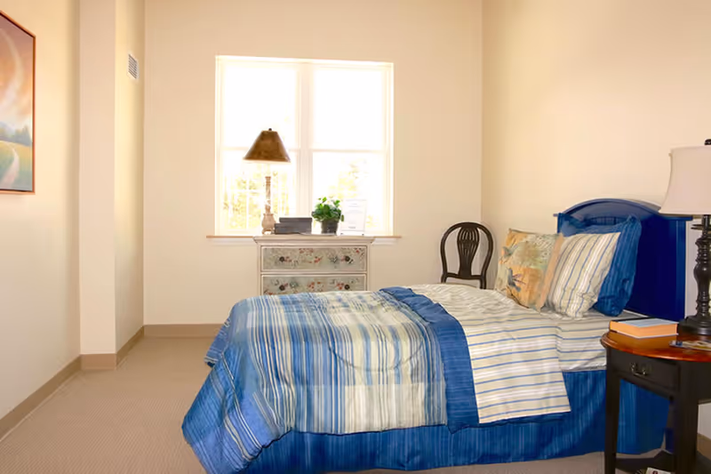 A tidy bedroom with a blue-striped bed, nightstand with a lamp, a decorative dresser under a window, and a chair.
