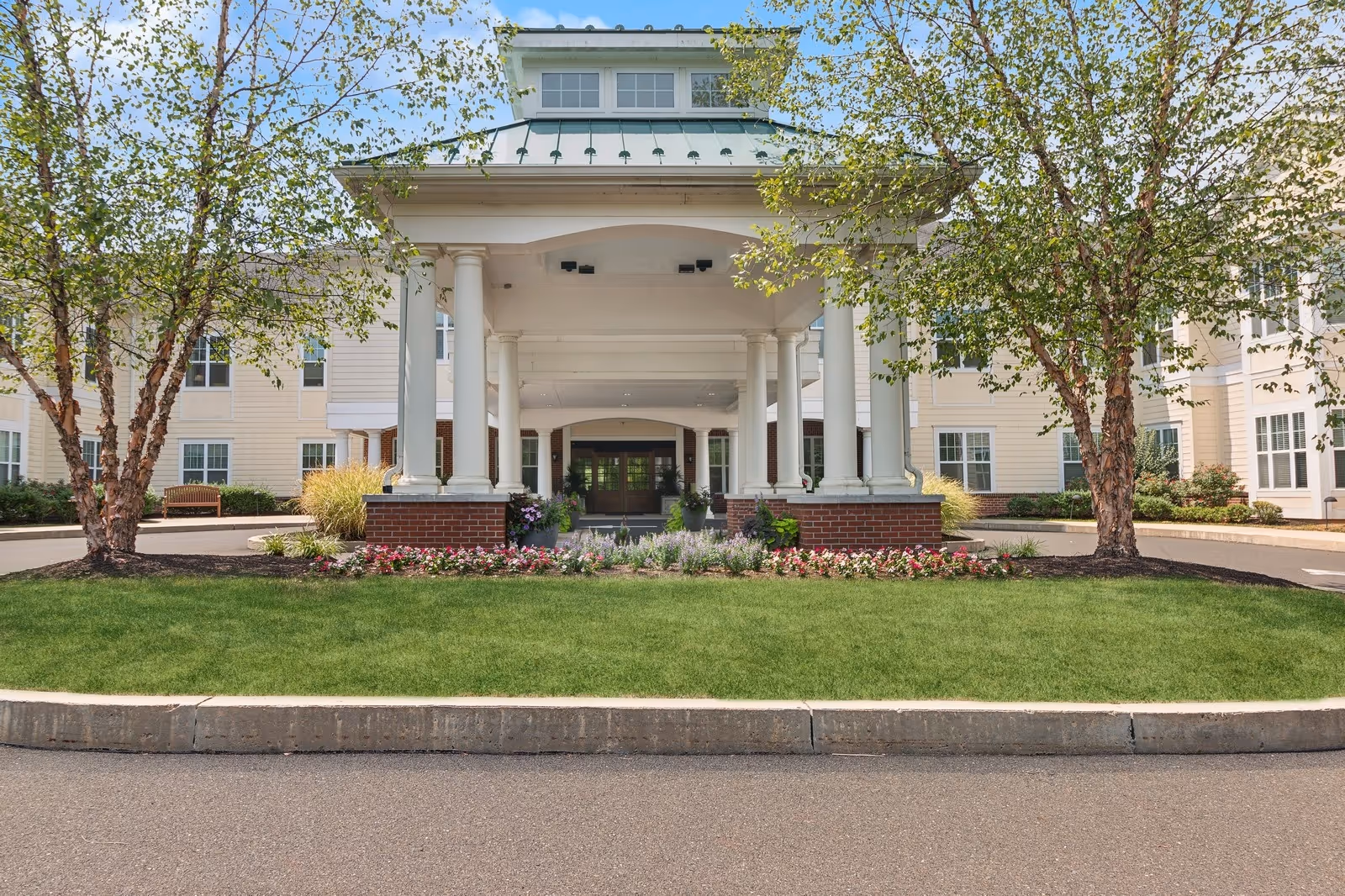 Front entrance of a senior living facility with a covered driveway supported by white columns, surrounded by green grass, flower beds, and two trees on either side. The building is light-colored with multiple windows and a clear blue sky above.