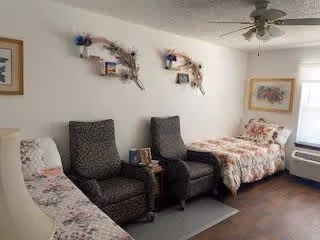 A cozy room with two dark upholstered armchairs placed side by side between a floral-patterned sofa and a bed with a floral bedspread. The room has light-colored walls decorated with framed artwork and wall-mounted floral arrangements. A ceiling fan is visible above, and a window with blinds lets in natural light. The floor is dark wood, and there is a small side table between the armchairs.