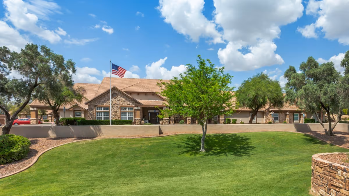 Exterior view of a single-story senior living facility building with a stone facade and tan roof, surrounded by green grass, trees, and landscaping under a partly cloudy blue sky. An American flag is flying on a flagpole in front of the building.