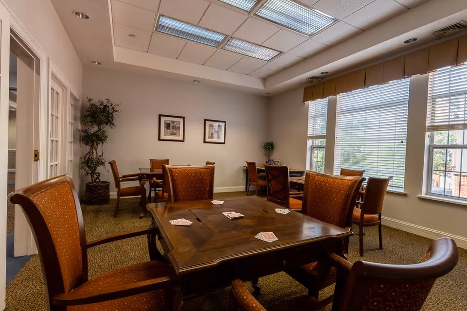 A well-lit room with multiple wooden tables and cushioned chairs arranged for group activities. Playing cards are spread out on the table in the foreground. The room has large windows with blinds, framed pictures on the wall, and a potted plant in the corner.