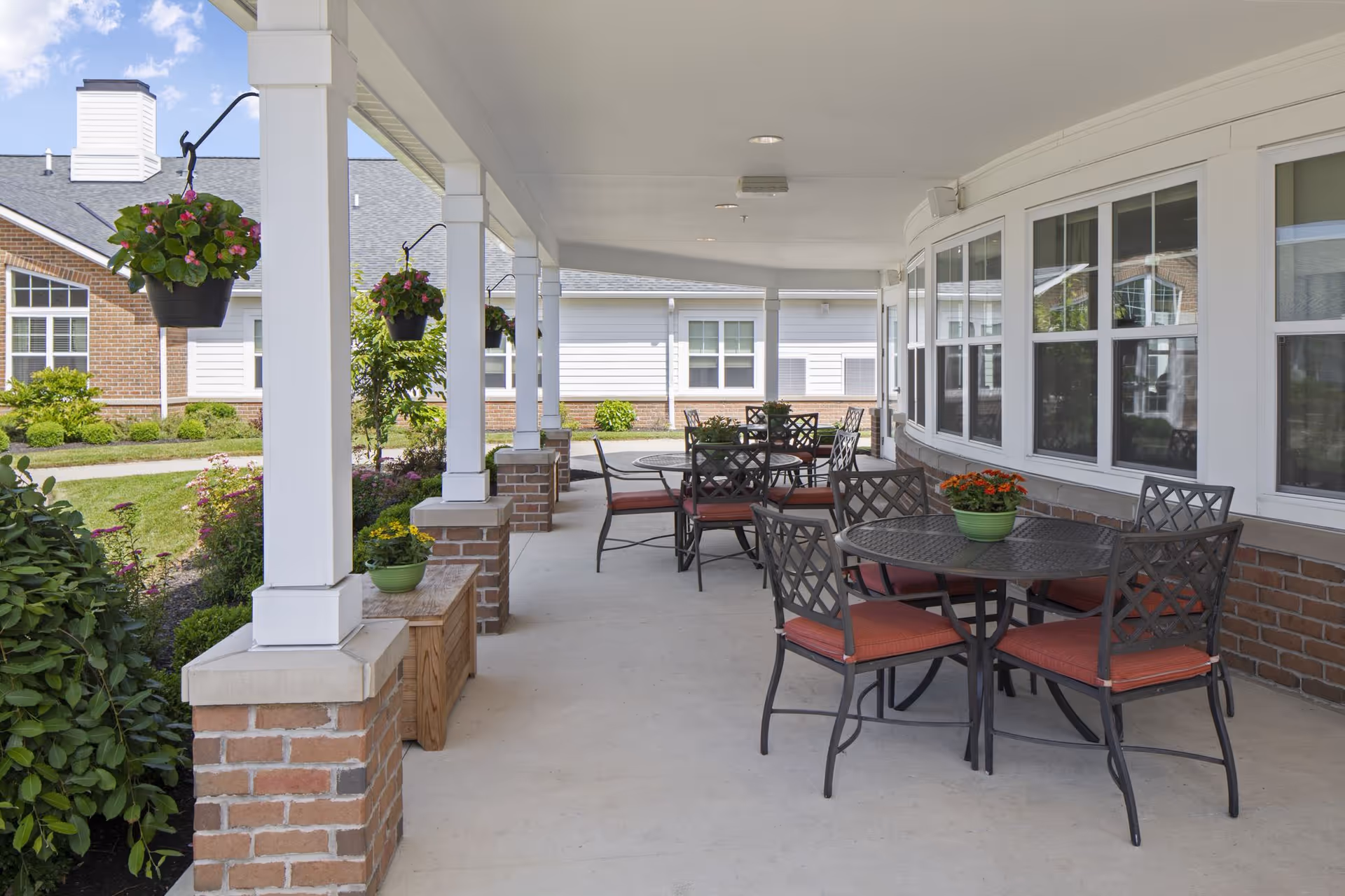 Covered outdoor patio area with several round metal tables and chairs with red cushions. Hanging flower pots and potted plants decorate the space. The patio is adjacent to a brick and white siding building with multiple windows.