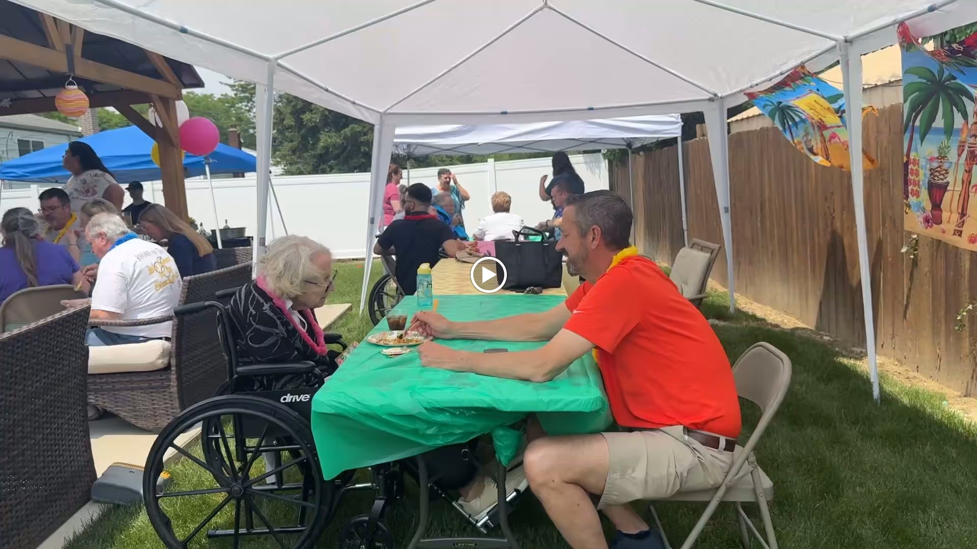 An outdoor gathering at Stratford House with elderly residents and staff seated under white canopy tents. One elderly woman in a wheelchair is sitting at a table covered with a green tablecloth, interacting with a man in a red shirt and beige shorts. Other people are seated and standing in the background, enjoying the event on a grassy lawn with a wooden fence and festive decorations.