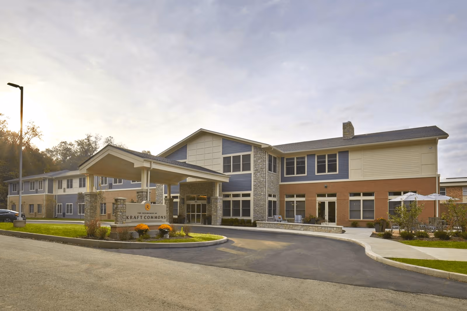 Exterior view of a two-story senior living facility building with a covered entrance and a sign that reads 'The Residences at Kraft Commons'. The building features a mix of stone, brick, and siding with multiple windows and a small outdoor seating area with umbrellas on the right side.