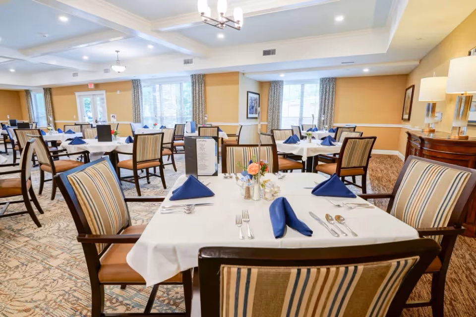Spacious dining room with neatly set tables, striped chairs, white tablecloths, and blue napkins.