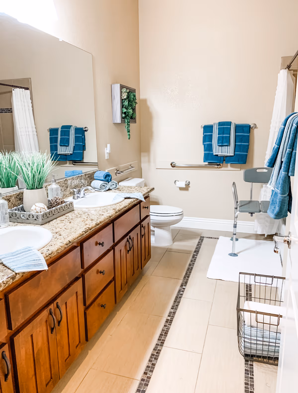 A clean and well-lit bathroom featuring a double sink vanity with granite countertop and wooden cabinets. There are decorative plants and neatly folded towels on the counter. The bathroom includes a toilet with grab bars, a shower area with a white curtain, and a shower chair. Blue towels hang on the wall and a basket with towels is on the floor.