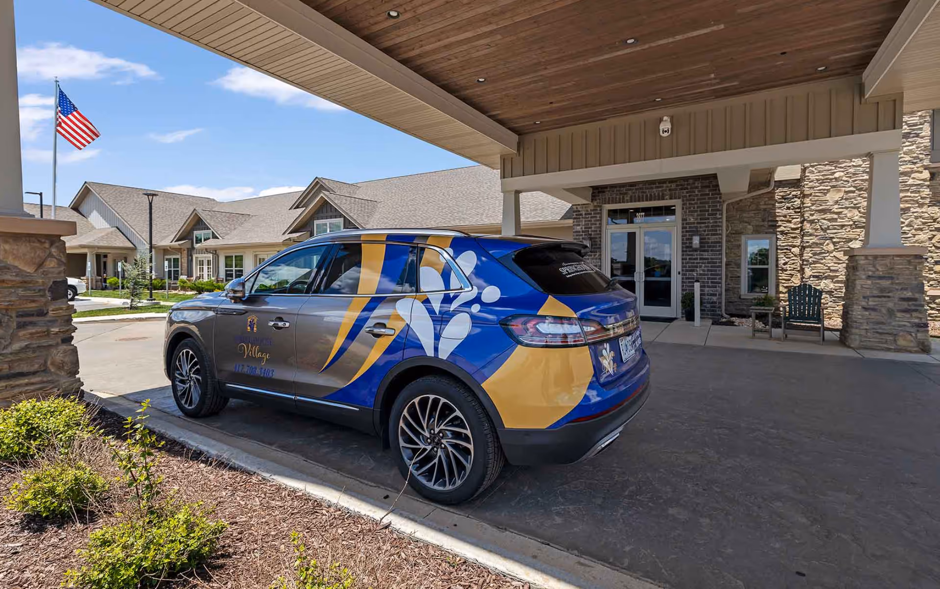 A vehicle with Springhouse Village branding parked under a covered entrance of a senior living facility. The building has stone and brick exterior walls, with a glass door entrance. An American flag is visible in the background on a flagpole.