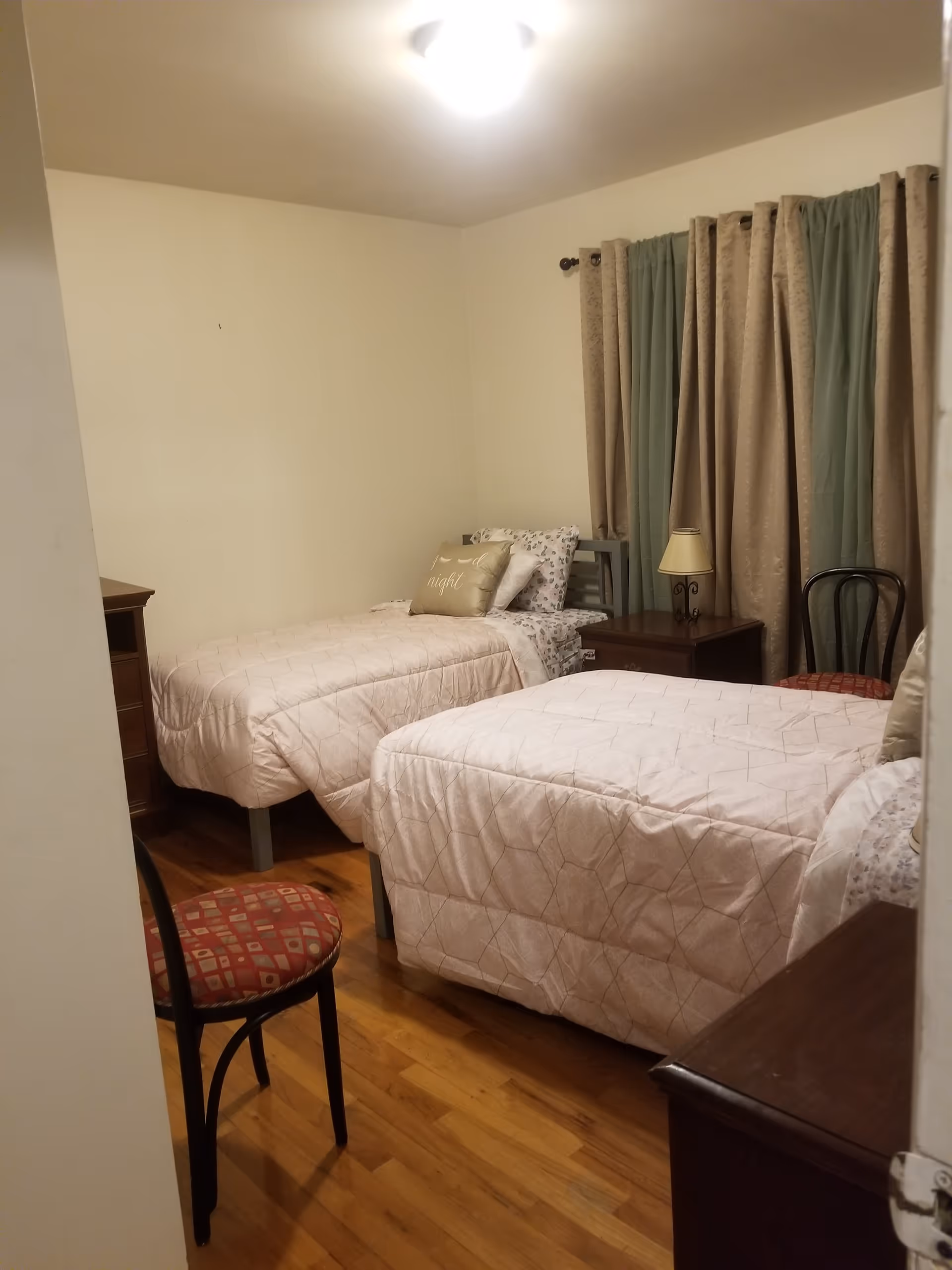 A small bedroom with two twin beds covered in light pink quilts. Each bed has pillows, one with a decorative pillow that says 'good night'. There is a wooden nightstand between the beds with a small lamp on it. The room has hardwood floors, beige walls, and a window with green and beige curtains. Two chairs with red patterned cushions are also visible in the room.