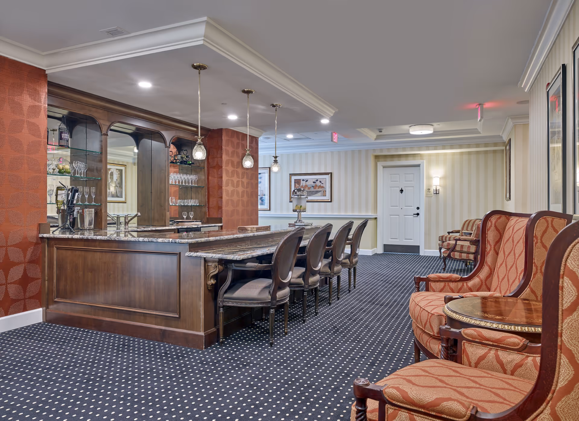 Interior view of a bar area with a polished wooden counter, high-backed chairs, and decorative lighting. The walls are adorned with a patterned design, and there are glasses displayed on shelves behind the bar.