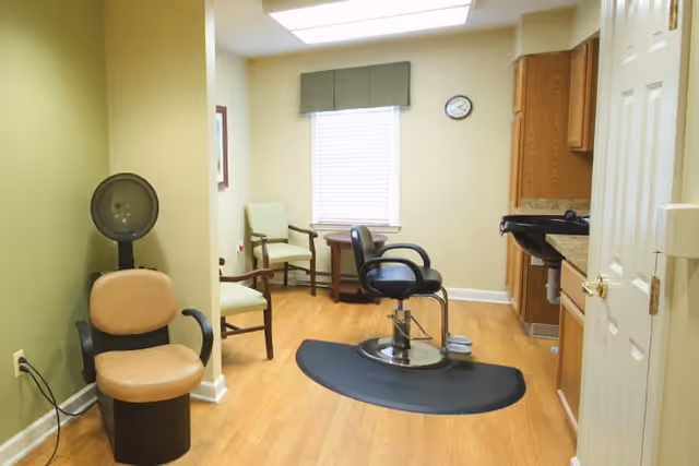 Interior view of a salon area in a senior living facility with a hair dryer chair on the left, a barber chair in the center on a black mat, wooden flooring, light green and beige walls, a window with a valance, two green chairs and a small round table near the window, and wooden cabinets with a sink on the right.