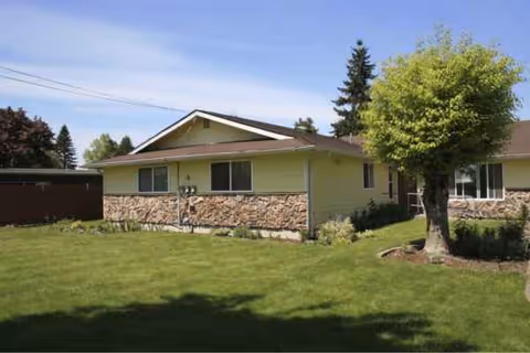 Single-story building with stone front, light yellow siding, green lawn and trees.