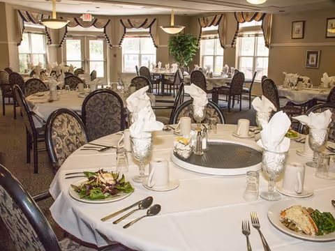 A dining room with round tables covered in white tablecloths, set with plates of food, silverware, glasses, and napkins folded in an elegant style. The room has large windows with valances, patterned chairs, and soft lighting fixtures hanging from the ceiling.