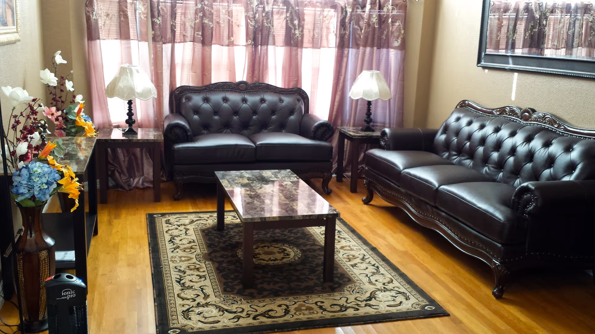 A living room with two dark brown leather tufted sofas, a marble-top coffee table on a patterned area rug, two side tables each with a lamp, a large mirror on the wall, and a vase with colorful artificial flowers. The room has wooden flooring and purple curtains covering the windows.