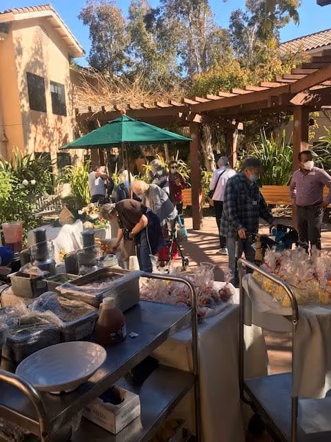 Outdoor courtyard area at a senior living facility with several people wearing masks gathered around tables and carts filled with various food items and supplies. There is a green umbrella providing shade and a wooden pergola overhead. The background shows trees and parts of the building with a tiled roof.