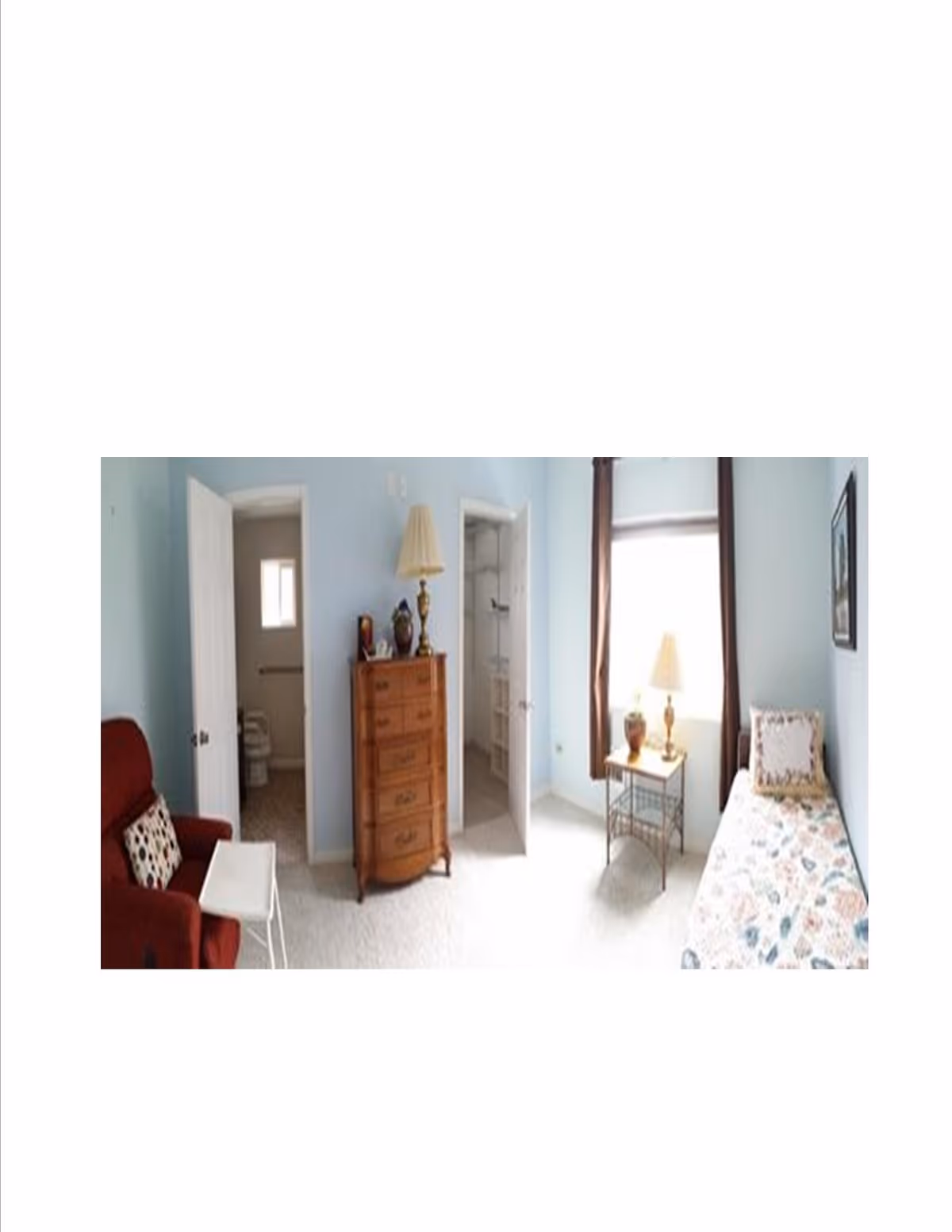 A small, cozy bedroom with light blue walls featuring a single bed with a floral bedspread, a wooden chest of drawers with a lamp and decorative items on top, a red armchair with a patterned pillow, a small white side table, and a window with brown curtains letting in natural light. Two open doors reveal a bathroom and a shower area.