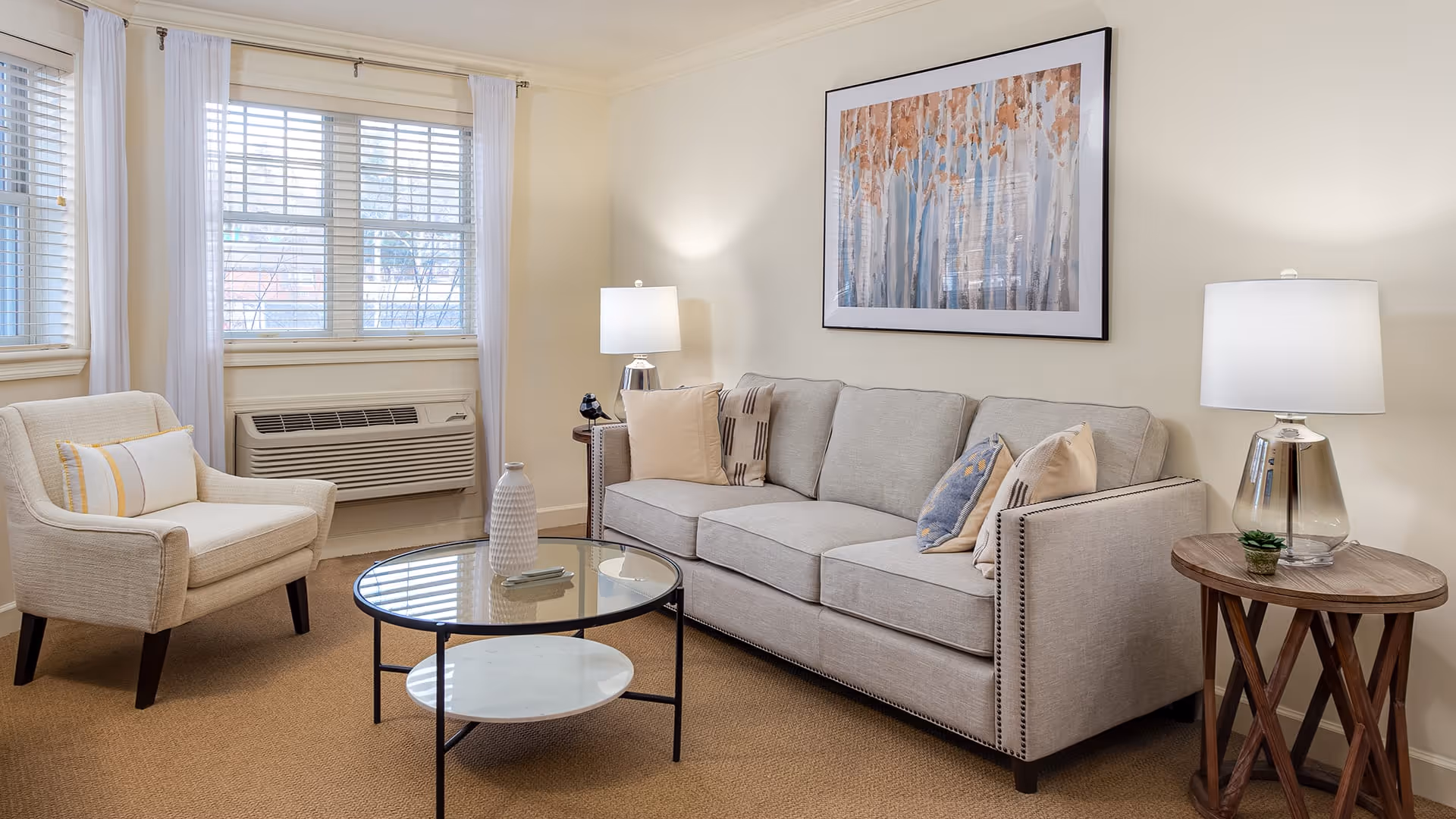 A cozy living room with a beige sofa adorned with multiple pillows, a matching armchair, a round glass coffee table with a decorative vase, and two side tables each with a lamp. The room has large windows with white curtains and a framed artwork of trees on the wall.