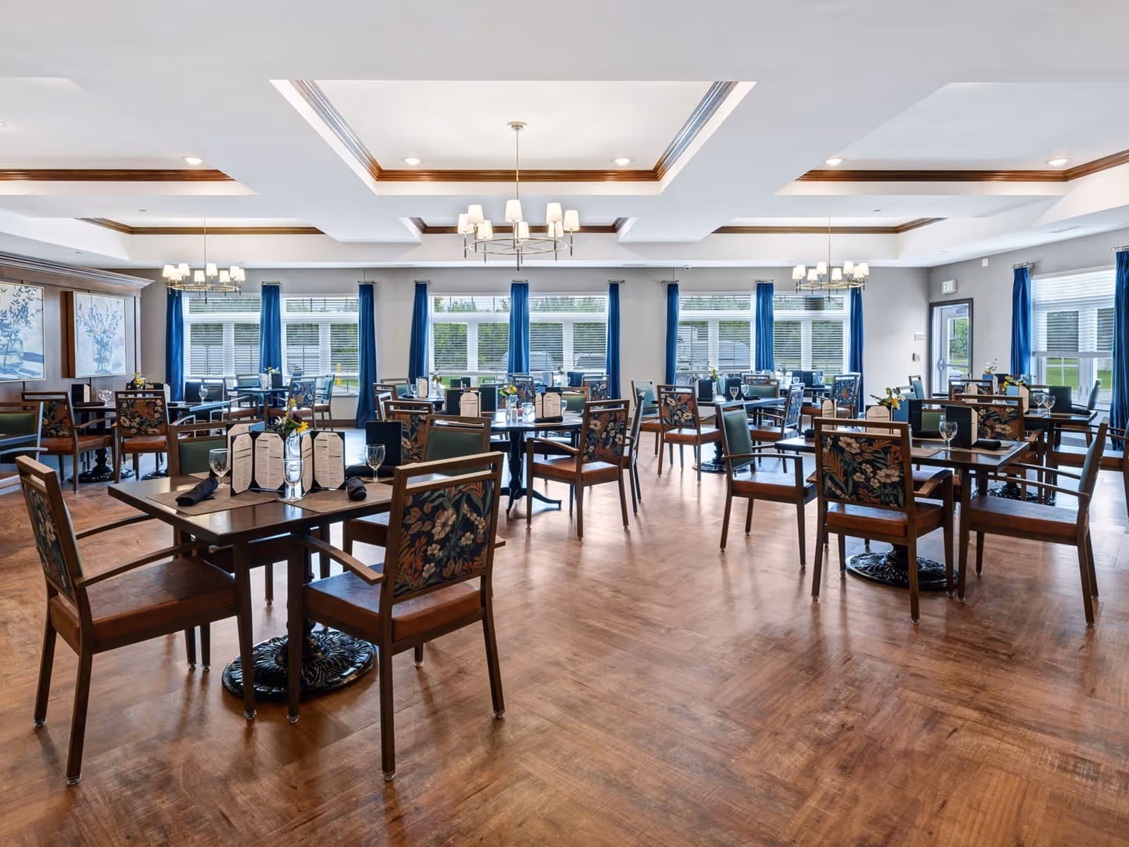 A spacious dining room with multiple wooden tables and chairs arranged neatly. Each table has menus, glassware, and napkins. The room features large windows with blue curtains, wooden flooring, and elegant ceiling light fixtures.