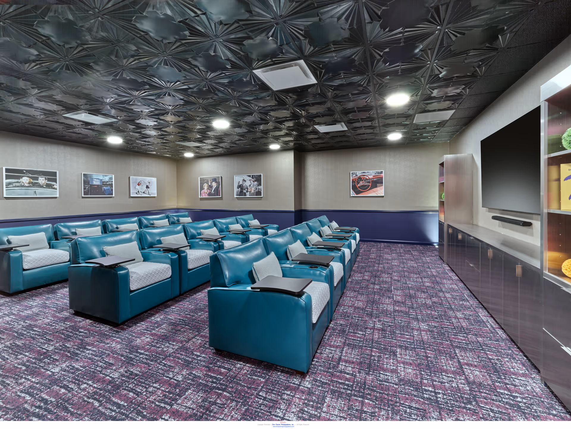 A media/theater room with teal reclining seats arranged in rows facing a large wall-mounted TV and framed pictures on the walls.
