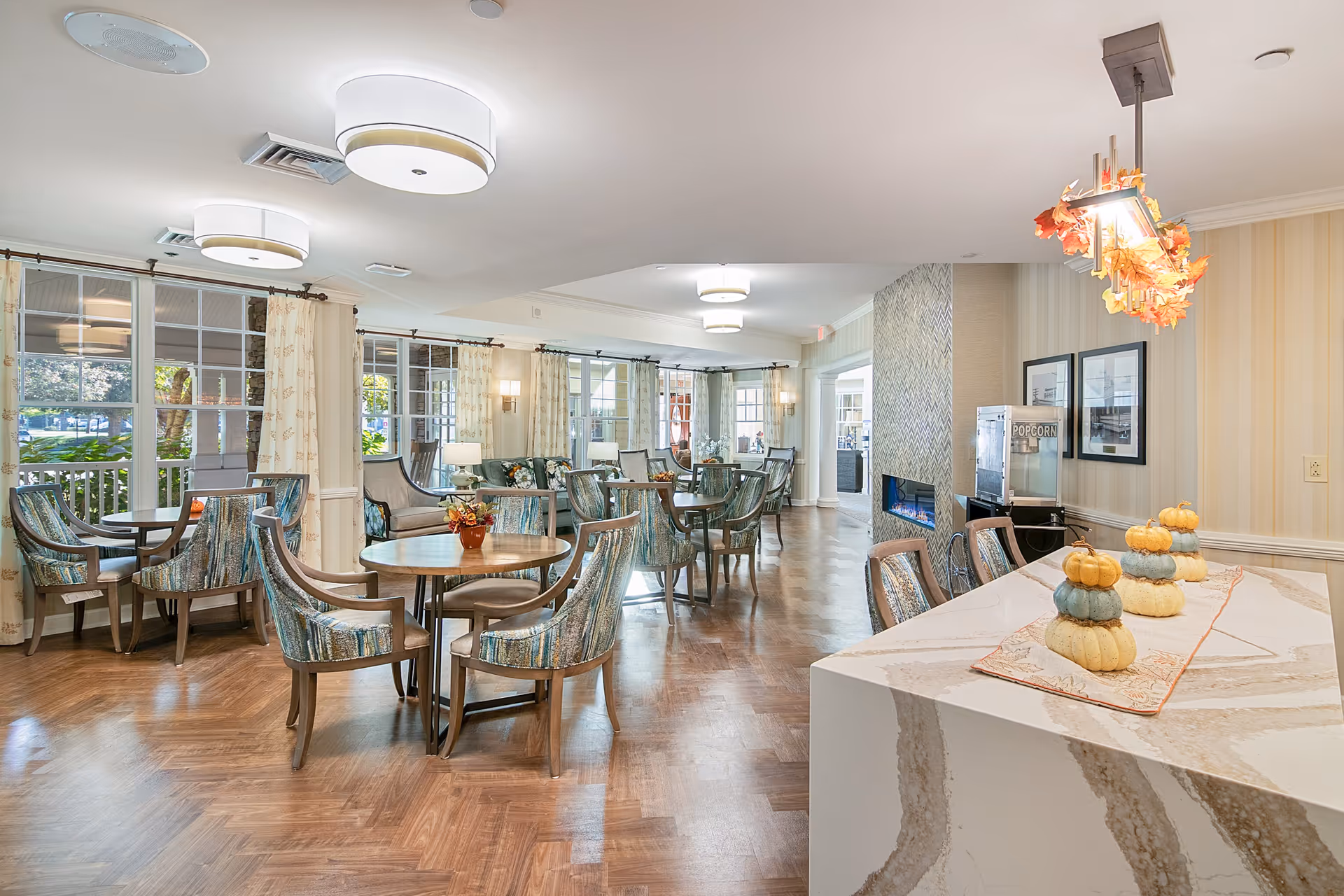 Bright and spacious dining area with multiple round tables and upholstered chairs. Large windows with curtains allow natural light to fill the room. A modern fireplace is visible on the right side, next to a popcorn machine. A marble countertop with decorative pumpkins and a hanging light fixture with autumn leaves is in the foreground.