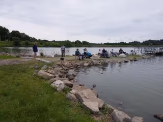 Several people sitting and standing on a rocky shoreline next to a calm body of water with grassy areas and trees in the background under a cloudy sky.