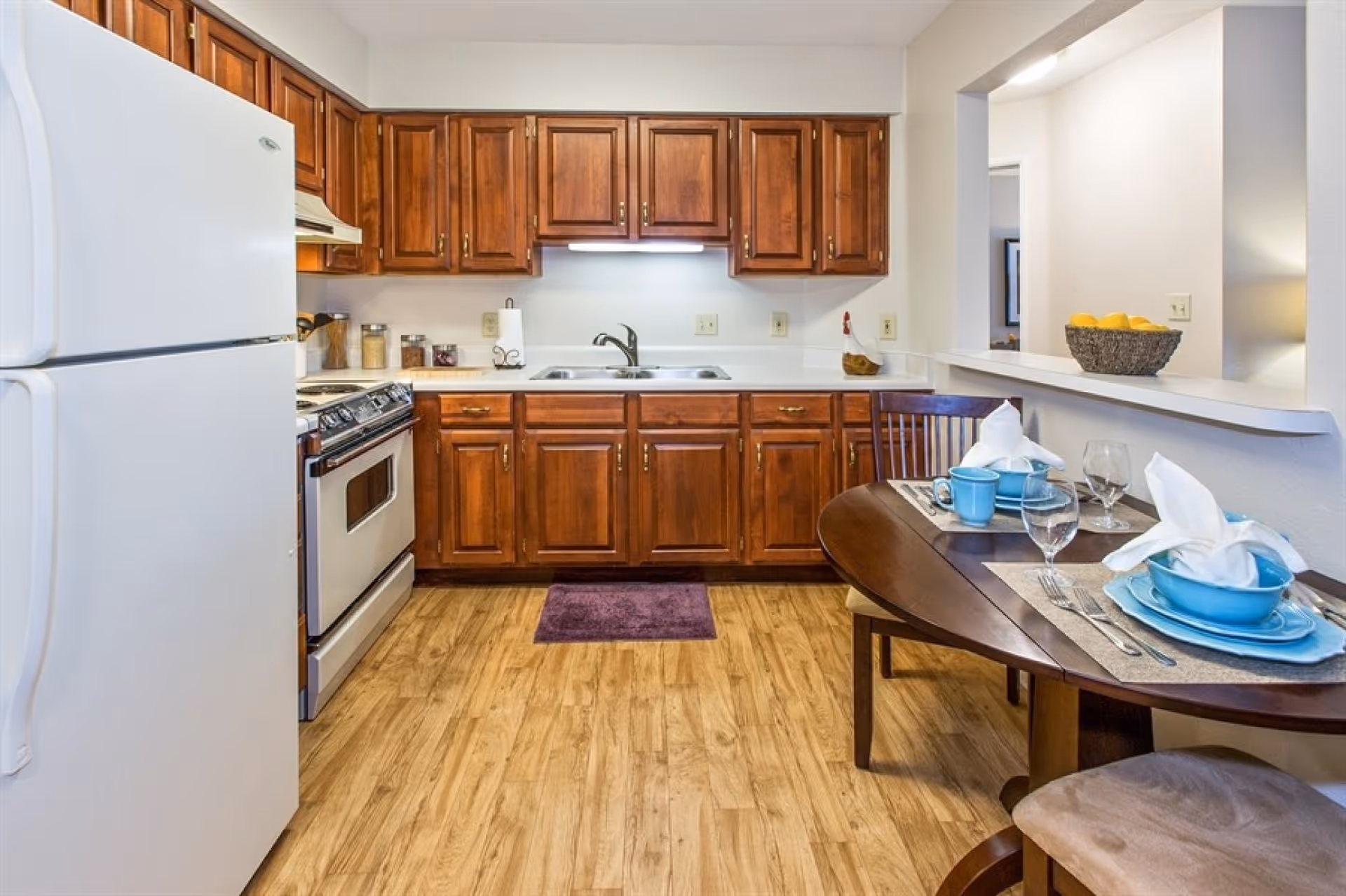 A kitchen with wooden cabinets, a white refrigerator, a stove, and a double sink. There is a small dining table set for two with blue dishes, white napkins, and glassware. The floor is wood, and there is a purple rug in front of the sink. A basket of lemons is on the counter near the dining area.