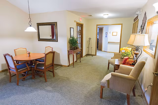 A cozy interior common area with a round wooden table and four chairs on the left, a hallway with doors and a small table with a plant in the center, and two armchairs with a side table and lamp on the right. The walls are light-colored and there is a framed mirror and artwork on the walls.