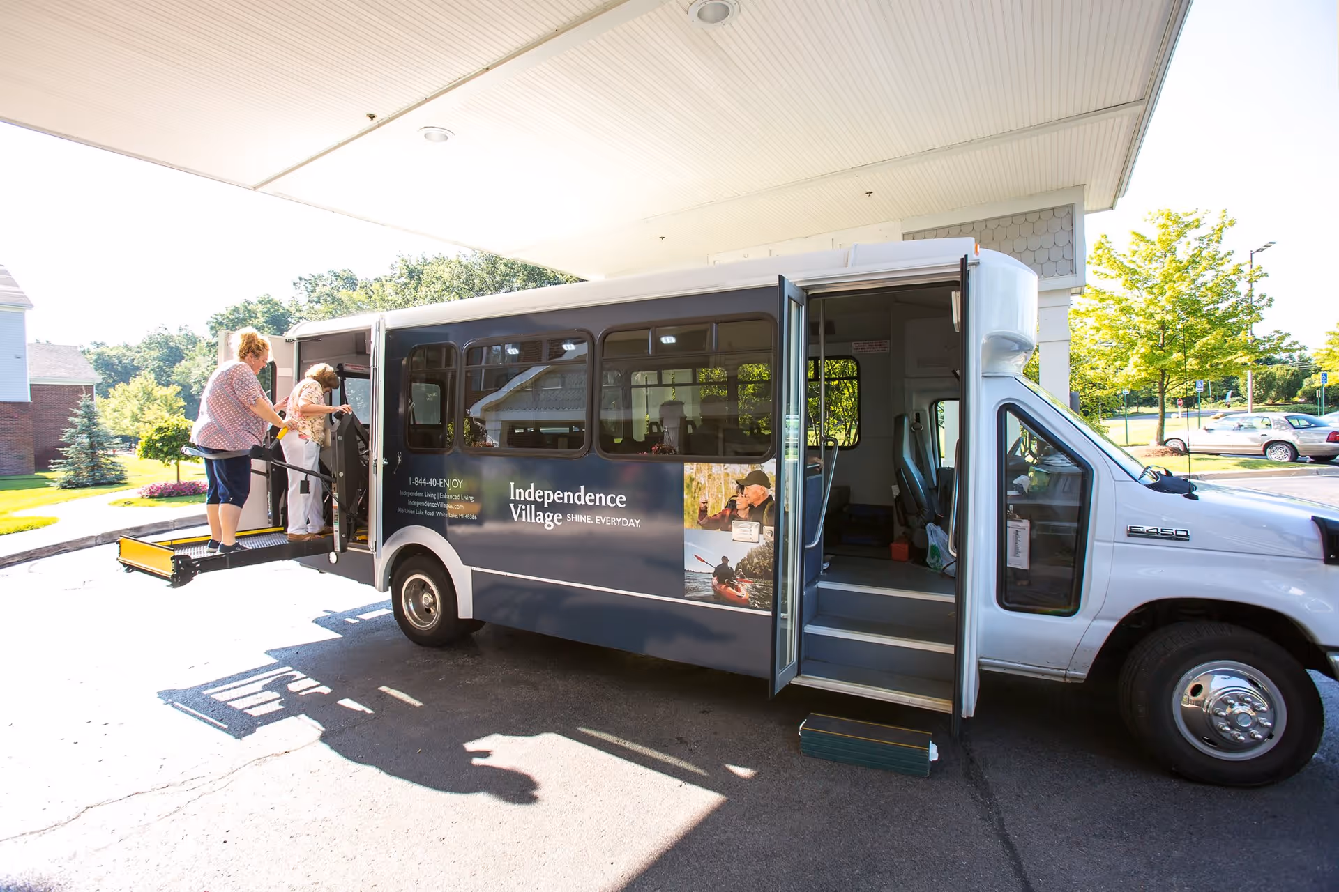 A shuttle bus from Independence Village of White Lake is parked under a covered area with its side door open. Two elderly women are using a wheelchair lift to board the bus. The bus is white and dark blue with the Independence Village logo and contact information on the side. The background shows a sunny day with trees, grass, and parked cars.