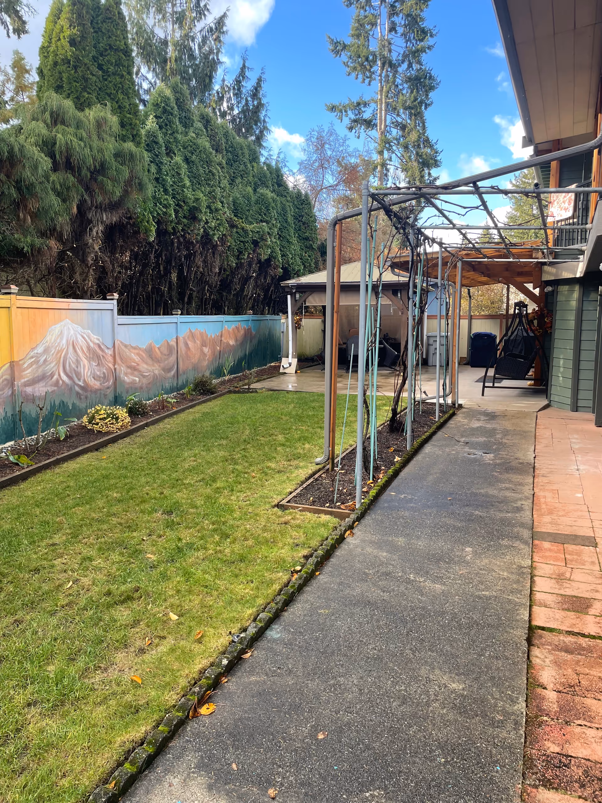 Outdoor area of a senior living facility with a paved walkway alongside a green lawn bordered by a garden bed. There is a mural of mountains painted on the fence to the left, tall evergreen trees behind the fence, and a covered patio area with seating and a swing on the right side near the building.