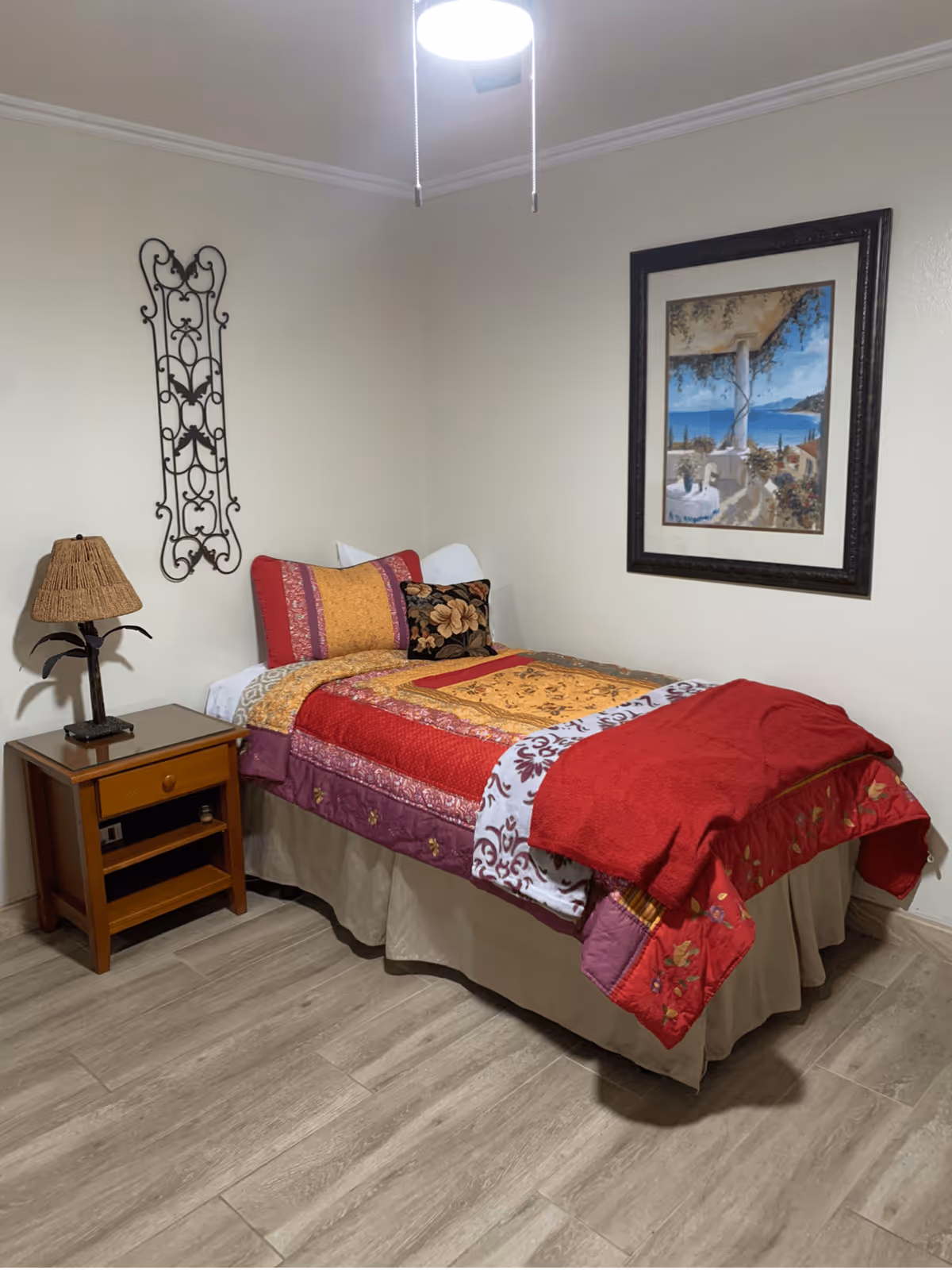 A small bedroom with a single bed covered in a colorful quilt with red, orange, and purple patterns. There is a wooden nightstand with a lamp featuring a woven lampshade and a decorative metal wall hanging above it. A framed painting of a seaside scene hangs on the wall above the bed. The room has light-colored walls and wood-look flooring.